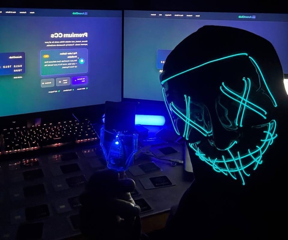 Person wearing a neon cyan stitched mask at gaming setup with dual monitors displaying code